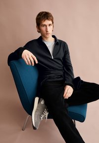 Dunkelgestreifter Reißverschluss-Pullover über einem grauen Hemd, schwarze Cordhosen, graue Turnschuhe mit weißen Akzenten, sitzt auf einem blauen Stuhl vor einem beigen Hintergrund.