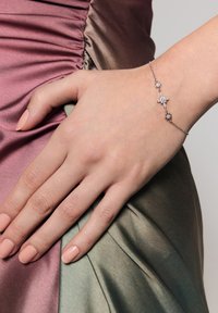 Srebrna bransoleta z pięcioma akcentami w kształcie gwiazd, zdobiona przezroczystymi kamieniami szlachetnymi. Tło zawiera satynową tkaninę w odcieniach różu i zieleni.