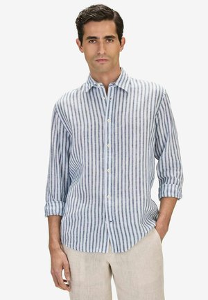 Mand iført en lyseblå og hvide vertikale stribede skjorte med knapper, ærmerne rullet op, kombineret med beige bukser, stående mod en hvid baggrund.