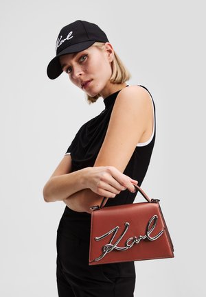 Borsa a mano in pelle marrone con una forma strutturata, dotata di un logo metallico argento "Karl". Il modello indossa un cappellino nero e una maglietta.