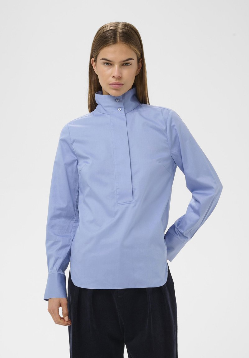 Chemise bleu clair avec col, patte de boutonnage à l'avant, manches longues et coupe cintrée. Tissu en coton lisse avec un léger éclat et des détails à boutons.