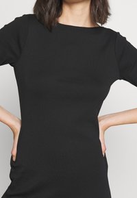 Žena v těsném černém topu s dlouhým rukávem, s rukama položenýma na bocích, před odlehčeným světlým pozadím.