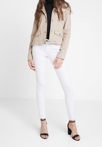 Kvinna iklädd beige knappad jacka, svart polotröja, vita smala jeans och svarta högklackade sandaler med vristband, stående mot vit bakgrund.