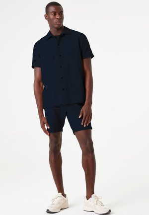 Camisa de manga corta de color navy y pantalones cortos a juego, fabricados con un tejido texturizado. Se lleva con zapatillas deportivas color crema, de pie sobre un fondo simple.