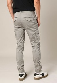 Man in een lichtgrijze cargobroek met klepzakken, zwart-witte sneakers en een zwart shirt, staande tegen een effen achtergrond.