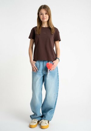 Młoda kobieta stojąca, ubrana w brązowy t-shirt, jasnoniebieskie luźne jeansy z czerwonym breloczkiem w kształcie serca i białe sneakersy na platformie.