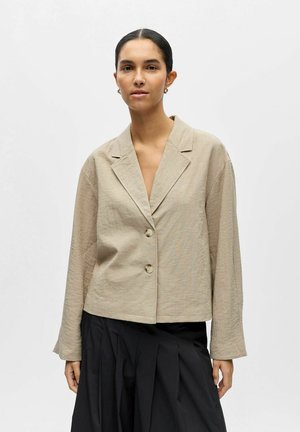 Frau mit dunklen Haaren, die einen beige geknöpften Blazer und einen plissierten schwarzen Rock trägt und vor einem schlichten hellen Hintergrund steht.