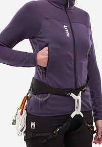 Personne portant une veste Millet violette et un harnais d'escalade noir avec des mousquetons et une corde attachés, la main dans la poche de la veste.
