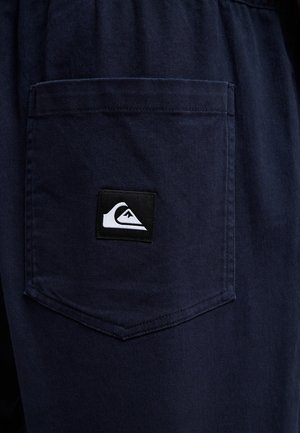 Blå stoffshorts med en baklomme som har en svart-hvitt logopatch. Materialet ser slitesterkt ut med en glatt tekstur.