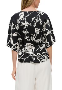 Zwarte bloemenblouse met witte abstracte patronen, korte mouwen en een ontspannen pasvorm, gecombineerd met lichtgekleurde broeken.
