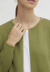 Un cardigan vert texturé en tissu tricoté, avec un col rond et une conception à ouverture sur le devant, superposé à une chemise blanche en dessous.