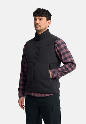 Man wearing black zip-up vest over purple and black checkered shirt with hand in vest pocket against plain background.