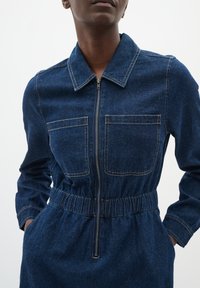 Denim overall met lange mouwen, kraag, elastische taille, twee borstzakken en een ritssluiting aan de voorkant in diepblauw. Gladde textuur.