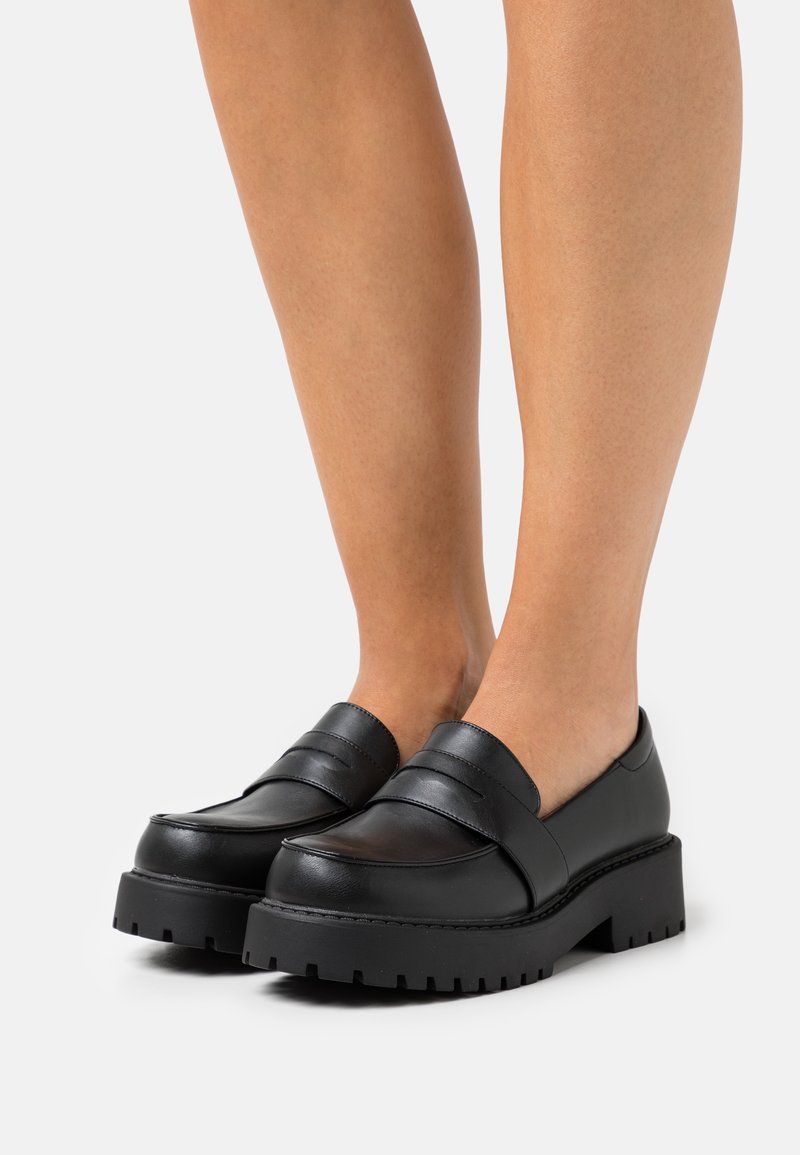 Mocassins noirs épais avec semelles crantées épaisses portés pieds nus sur un fond clair uni.