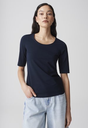Vrouw met lang donker haar, gekleed in een navyblauw T-shirt met korte mouwen en lichtblauwe spijkerbroek, staand tegen een effen lichte achtergrond.