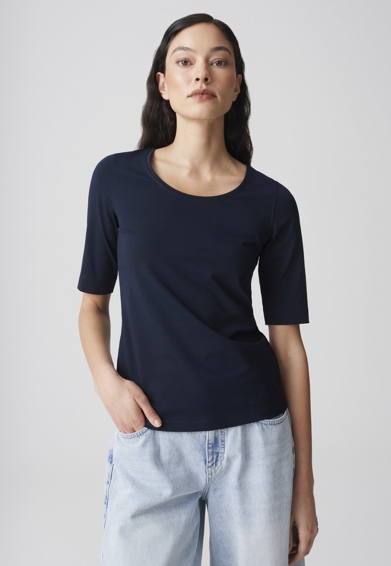 Vrouw met lang donker haar, gekleed in een navyblauw T-shirt met korte mouwen en lichtblauwe spijkerbroek, staand tegen een effen lichte achtergrond.