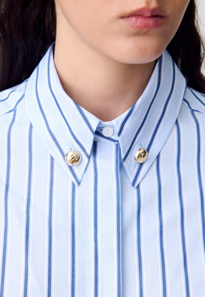 Primer plano del cuello de una persona que lleva una camisa azul clara con rayas verticales blancas y azules y dos botones decorativos dorados.