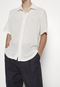 Camisa de botones de manga corta blanca en un tejido de punto texturizado, que presenta un cuello, un patrón de tejido abierto y un corte relajado.