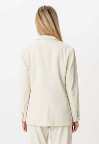 En blondhåret person, der vender bort fra kameraet, iført en cremegul jakkesæt og matchende bukser mod en hvid baggrund.