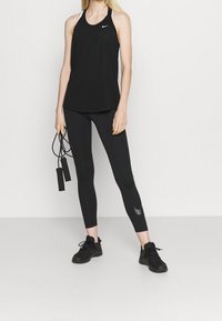 Haut de sport noir sans manches avec accents blancs, associé à un legging noir arborant un petit logo, tenant une corde à sauter noire. Baskets noires.