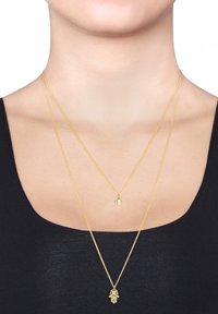 Tre lagdelte guldhalskæder, der har en hamsa-amulet, et øje design og delikate kædetræk, præsenteret på en sort top mod en hvid baggrund.