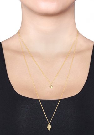 Tre lagdelte guldhalskæder, der har en hamsa-amulet, et øje design og delikate kædetræk, præsenteret på en sort top mod en hvid baggrund.