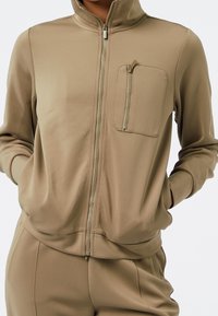 Beige idrottsjacka med dragkedja, tillverkad av ett slätt tyg, med hög krage och en dragkedjeförsedd bröstficka. Modellen poserar med händerna i fickorna.