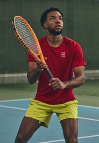 Rød treningsskjorte og neongrønne shorts. Mannen holder en oransje tenniskølle, klar til å spille på en blå bane med teksturert overflate.