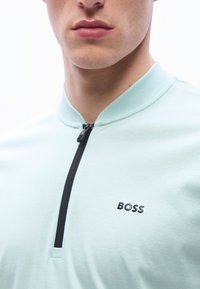 Κοντινή λήψη ενός ατόμου που φοράει ανοιχτό μπλε τοπ με φερμουάρ στο κέντρο και το λογότυπο "BOSS"  στο στήθος, με ορατά μόνο τα χείλη και το κάτω μέρος του προσώπου.