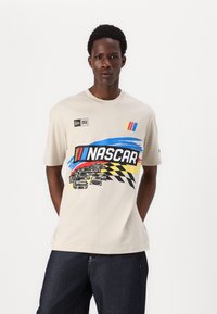 T-shirt en coton beige avec un graphique NASCAR présentant des voitures de course multicolores, des rayures et un motif à damier sur le devant.