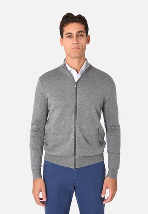 Cardigan grigio con zip, colletto alto, texture a maglia, due tasche frontali e polsini a coste. Abbinato con una camicia bianca con bottoni e pantaloni blu.
