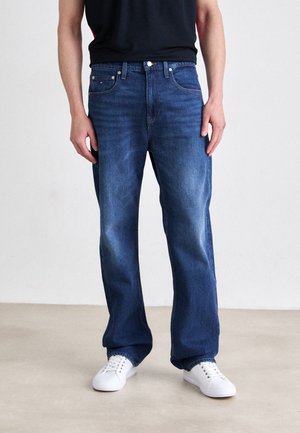 Mann trägt locker sitzende blaue Jeans mit schwarzem Hemd und weißen Sneakers, steht auf hellem Boden vor einer schlichten Wand.