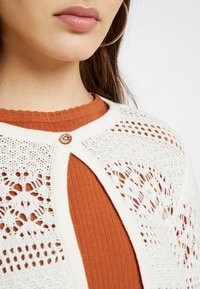 Cardigan bianco lavorato a maglia con motivo in pizzo, caratterizzato da un bottone di legno e un top a coste color ruggine sotto. Dettagli a maglia texturizzati evidenti.