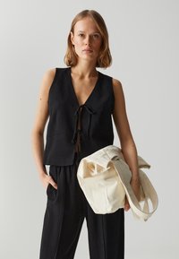 Top senza maniche in lino nero con scollatura a V profonda e due tasche frontali. Il modello tiene in mano una borsa tote in tela color crema.
