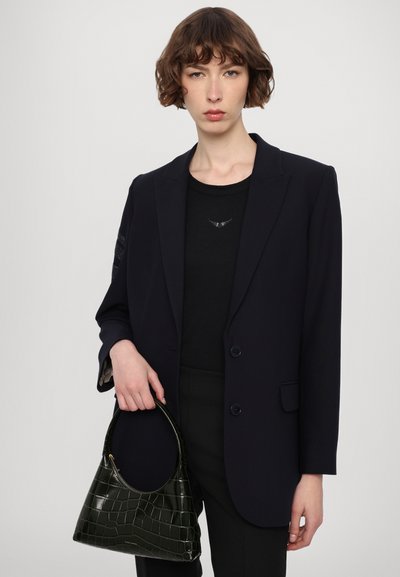 Blazer bleu marine foncé au design structuré, avec des fermetures à boutons, et une chemise noire arborant un petit emblème d'aigle. Le mannequin tient un sac à main en croco vert.