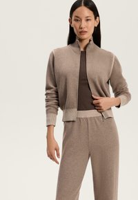 Rjava pletena jakna na zadrgo z visokim ovratnikom, rebrastimi manšetami in usklajenimi rjavimi hlačami; mehak material in sproščen kroj.