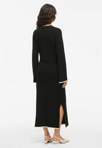 Černé šaty s dlouhým rukávem s žebrovanou strukturou, boční rozparkou a krémovým lemováním kolem výstřihu a rukávů. Nošeno s světlými botami.