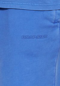 Gros plan sur un tissu bleu avec le texte en relief « DROPSIZE » près d’une couture et de la ceinture d’un vêtement, probablement un jogging ou un short.