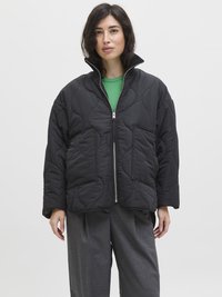 Veste rembourrée noire avec un col haut, présentant une texture matelassée et une fermeture éclair. Portée au-dessus d'un haut vert et d'un pantalon gris.