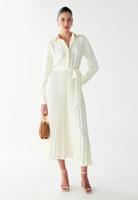 Helles Creme-farbenes, plissiertes Kleid mit langen Ärmeln, einem Kragen und einem Taillengürtel. Ergänzt mit einer kleinen braunen Tasche und weißen Riemchen-Absätzen.