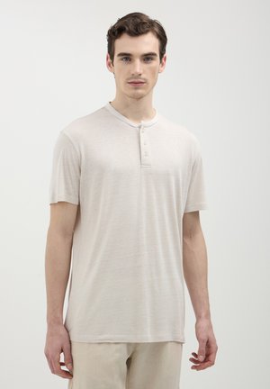 Jeune homme avec des cheveux bouclés courts portant une chemise henley à manches courtes beige clair et un pantalon assorti, se tenant devant un fond blanc uni.