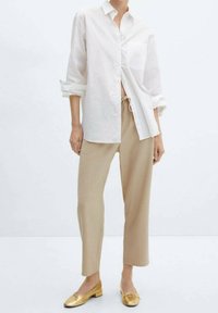 Weißes Hemd mit hochgekrempelten Ärmeln, beigefarbene, kürzere Hose und goldene Ballerinas. Glatte Texturen, lässige Passform und minimalistisches Design.