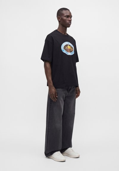 Man standing wearing a black oversized graphic t-shirt, wide-leg dark jeans, and white sneakers on a plain light background.