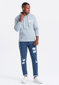 Hombre con una sudadera azul clara que tiene el texto "WILD", jeans azul desgastados y zapatillas blancas, de pie y mirando hacia un lado.