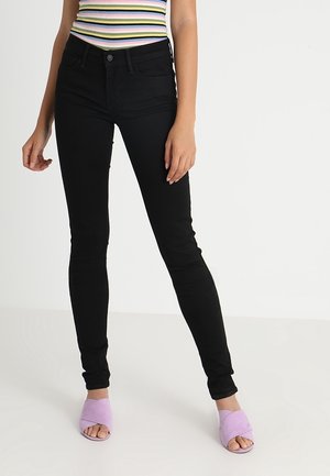 Femme portant un jean skinny noir, un haut sans manches rayé pastel et des sandales ouvertes violet clair, debout sur un sol blanc.