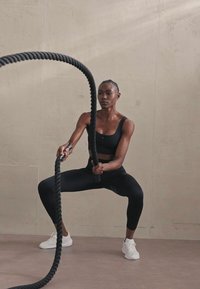 Melna sporta krūšturis un augsta jostasvieta legingi, kas tur resnu, teksturētu kaujas virvi. Fons ar gludām, gaišām sienām.