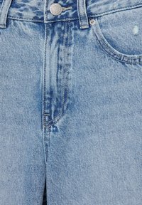 Jean en denim bleu clair, gros plan sur le devant montrant le bouton, la zone de la fermeture éclair, les passants de ceinture et la poche avant avec une légère usure du tissu.