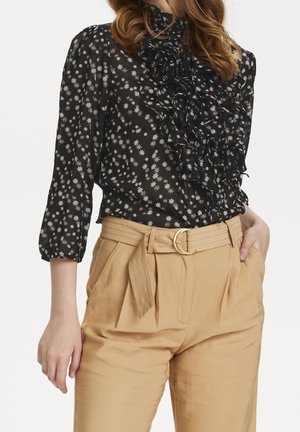 Blouse noire avec des motifs d'étoiles blanches, détails volantés au cou et manches trois quarts, associée à un pantalon beige taille haute.