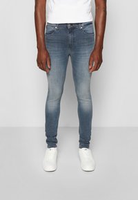 Úzké modré džíny z denimu s vybledlým odstínem, pětistranným designem, zipovým zapínáním a jemnými detaily šití. Spárovány s bílými sportovními botami.