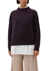 Femme portant un pull côtelé violet foncé sur une chemise blanche, associée à un pantalon large beige, se tenant devant un fond uni.